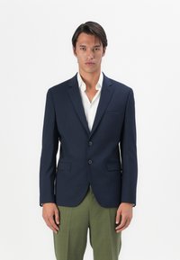 Marineblazer med en strukturert design, to knapper, slagkrage og to frontlommer, kombinert med en hvit skjorte og grønne bukser.