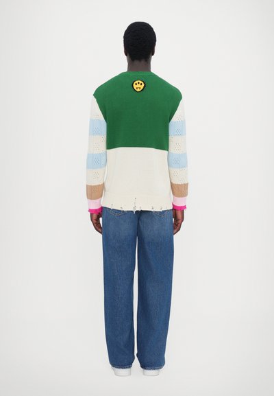 Striktrøje med grønne, blå og beige felter samt en smiley ansigt detalje på ryggen. Sammen med løse blå denimjeans.