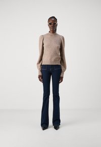 Brun strukturert genser med puffdetaljer på ermene, kombinert med mørkeblå utsvungne jeans og spisse svarte hæler. Tilbehøret inkluderer smale solbriller.