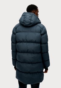 Abrigo largo acolchado en color navy con capucha, presenta amplios acolchados horizontales, una textura suave y puños elásticos. La vista trasera muestra la longitud y la forma.