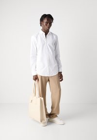 Hvid langærmet skjorte med knaplukning, parret med beige vide bukser. Modellen holder en lys beige tote taske og har hvide sneakers på.