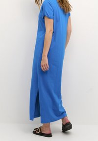 Robe bleue longue jusqu'aux chevilles avec une coupe décontractée, manches courtes retroussées, fente latérale et texture douce. Portée avec des sandales noires simples.