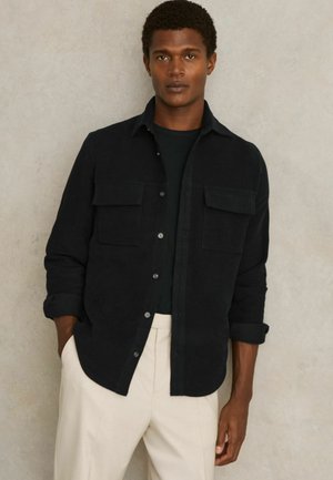 Camisa negra de botones en tejido suave con dos bolsillos en el pecho, llevada sobre una camiseta negra, combinada con pantalones crema. Textura suave, corte relajado.