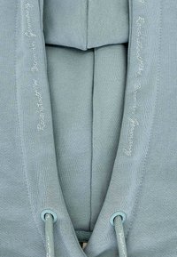 Felpa leggera e morbida di colore blu chiaro con cappuccio, dotata di un cordino al collo, tessuto testurizzato e scritte ricamate lungo i lati.