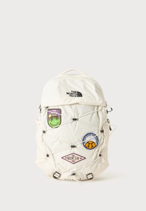 Hvid North Face rygsæk med tre farverige patches og sorte elastiske snore, der krydser foran.