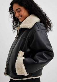 Sort læderjakke med cremet faux shearling krave og manchetter, med frontlynlås og sidelommer, løs pasform og struktureret overflade.