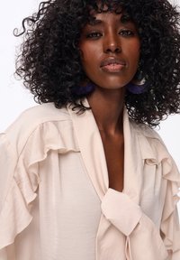 Kobieta z kręconymi włosami, nosząca jasnobeżową bluzkę z falbankami i duże fioletowe kolczyki z frędzlami na białym tle.