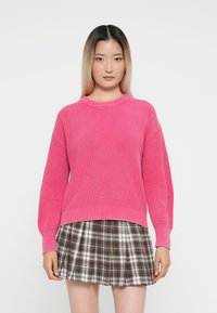 Suéter rosa canelado de manga longa e gola redonda, combinado com uma saia mini xadrez em tons de castanho e creme.