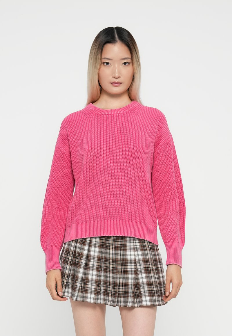 Suéter rosa canelado de manga longa e gola redonda, combinado com uma saia mini xadrez em tons de castanho e creme.