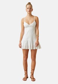 Vestido blanco sin mangas con un patrón texturizado, que presenta un dobladillo acampanado y tirantes ajustables. Combinado con sandalias marrones de tiras.