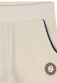 Pantaloni a coste di colore chiaro con una texture morbida, dotati di una toppa in pelle marrone e piping blu navy lungo le tasche.