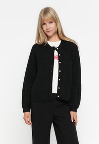 Cardigan noir en tricot torsadé avec un col rond, des boutons décorés de perles et des poignets côtelés, associé à un haut blanc.