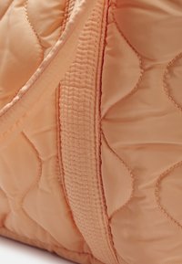 Sac matelassé de couleur pêche avec une finition lisse, des coutures détaillées et un accent texturé. Présente des formes douces et arrondies ainsi que des bretelles robustes.