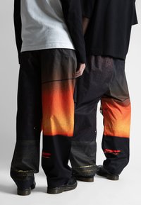Čierne nohavice s gradientným dizajnom, prechádzajúcim od oranžovej po tmavé odtiene, s voľným strihom, decentnými metalickými akcentmi a textúrovaným povrchom.