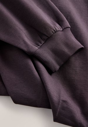 Lilla pusa tekstureeritud kangaga. Omadusteks on soonikkoes mansetid ja lõdvestunud lõige. Materjal on pehme ja veidi karvane.