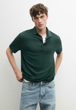 Jeune homme aux cheveux bruns bouclés portant un polo vert foncé à manches courtes sur un tee-shirt blanc et un jean bleu clair, debout devant un fond uni.