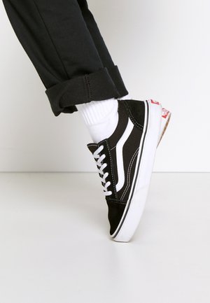 Baskets noir et blanc avec lacets blancs, portées avec des chaussettes blanches et un pantalon noir retroussé, montrant le côté et la semelle sur un fond uni.