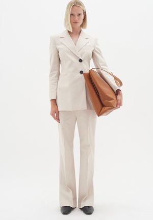 Beige op maat gemaakte pak met lange, rechte broek en een getailleerde blazer met twee zwarte knopen. Houdt een bruin leren tas vast.