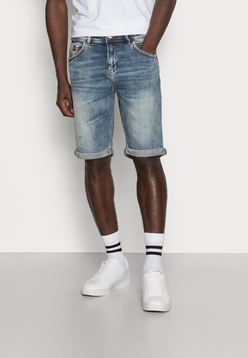 Shorts en denim bleu clair avec revers roulés, cinq poches et fermeture à bouton, associés à des baskets blanches et des chaussettes rayées.