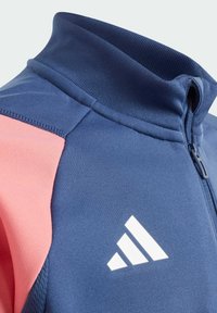 Rozpinana kurtka w granatowym kolorze z różowymi akcentami, ze żebrowanym kołnierzem i białymi detalami logo. Gładka struktura tkaniny w całej kurtce.