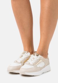 Beige and white sneaker with mesh and leather, gold accents, and round toe. Features white laces and a textured sole for grip.