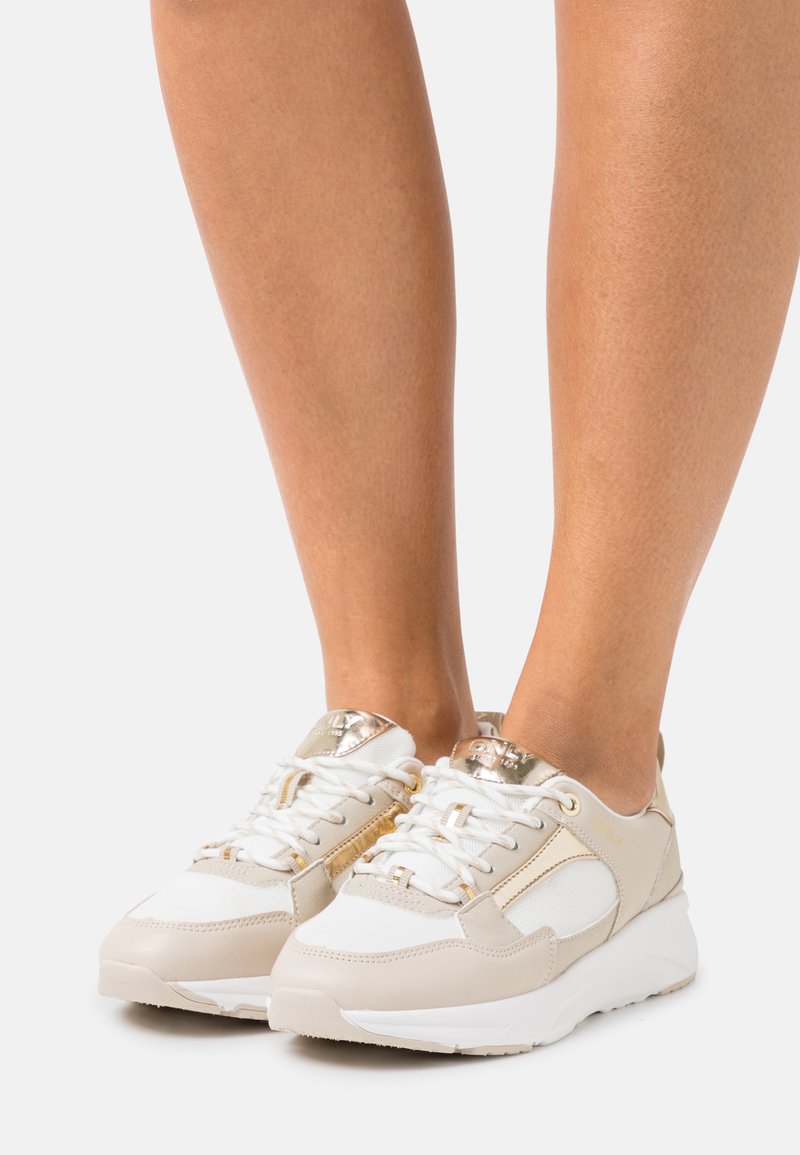 Beige and white sneaker with mesh and leather, gold accents, and round toe. Features white laces and a textured sole for grip.