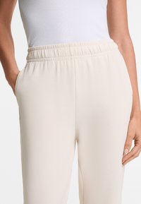 Light beige elastic-waist pants with side pockets, smooth texture, and a relaxed fit, paired with a white ribbed tank top.