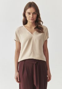 Beige satinate paita dengan leher V, lengan pendek, dan potongan longgar, dipadukan dengan celana pinggang tinggi burgundi tua yang dilengkapi dengan ikat pinggang.