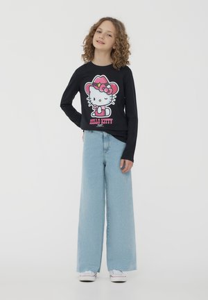 Czarna koszula z długim rękawem z kolorową grafiką Hello Kitty, w połączeniu z jasnoniebieskimi spodniami o szerokich nogawkach i białymi trampkami na jednolitym tle.