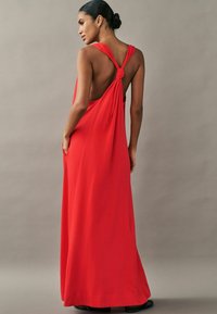 Červené maxi šaty bez rukávů s uzlem na zádech, v volném střihu a elegantním splývavém vzhledu. Materiál vypadá hladce a lehce.