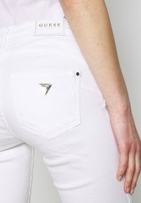 Bílé džíny vyrobené z denimu s trojúhelníkovým kovovým logem na zadní kapse, hladkým povrchem a přiléhavým střihem.