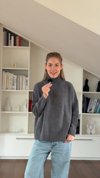 Suéter cinza de malha com gola alta e punhos e bainha canelados, combinado com jeans leves de corte solto em azul claro. Penteado simples e fundo neutro.