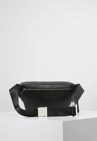 Musta kangaskotelo, jossa on sileä rakenne, vetoketjullinen etutasku ja säädettävä hihna, jossa on valkoinen solkiyksitys.