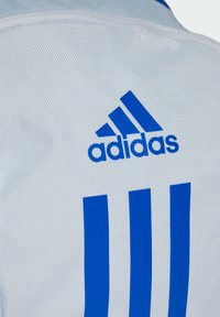 Chaqueta deportiva blanca con logo azul de adidas y tres franjas verticales azules. Hecha de tela texturizada, con detalles de costura.