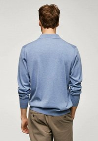 Uomo con capelli castano chiaro corti, che indossa un maglione a maniche lunghe blu chiaro e pantaloni beige, in piedi di spalle contro uno sfondo semplice.