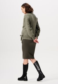 Olivgrüner Pullover mit Bindeband in der Taille, kombiniert mit einem gerippten olivgrünen Bleistiftrock und knielangen schwarzen Stiefeln. Glatte Textur insgesamt.