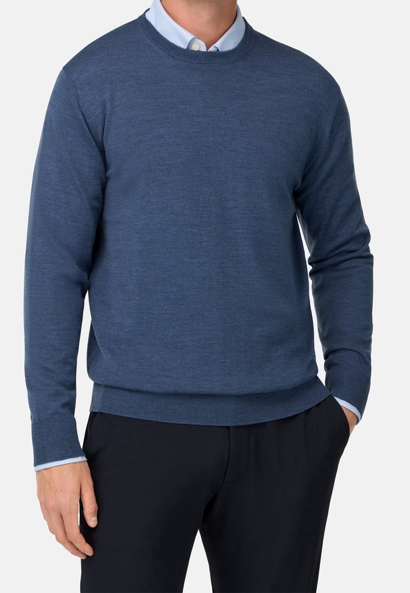 Homme portant un pull ras du cou bleu par-dessus une chemise à col bleu clair avec un pantalon noir, une main dans la poche, sur un fond uni.