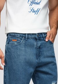 Vaqueros de mezclilla en azul intermedio con una textura ligeramente desgastada, bolsillos frontales planos y una etiqueta de cuero marrón en la cinturilla.