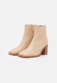 Beżowe botki do kostki wykonane z gładkiej skóry, z kwadratowym noskiem, bocznym zamkiem błyskawicznym oraz nałożonym drewnianym obcasem.