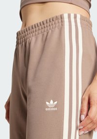 Bruna atletiska byxor med tre vita ränder, elastisk midja och liten vit logotyp på låret. Tillverkad av en texturerad väv.