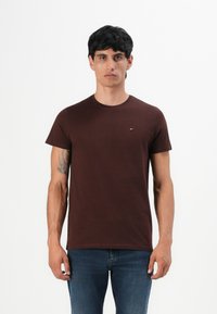 Ein Mann mit kurzen schwarzen Haaren trägt ein einfaches dunkelbraunes T-Shirt und blaue Jeans und steht vor einem weißen Hintergrund mit einem neutralen Ausdruck.