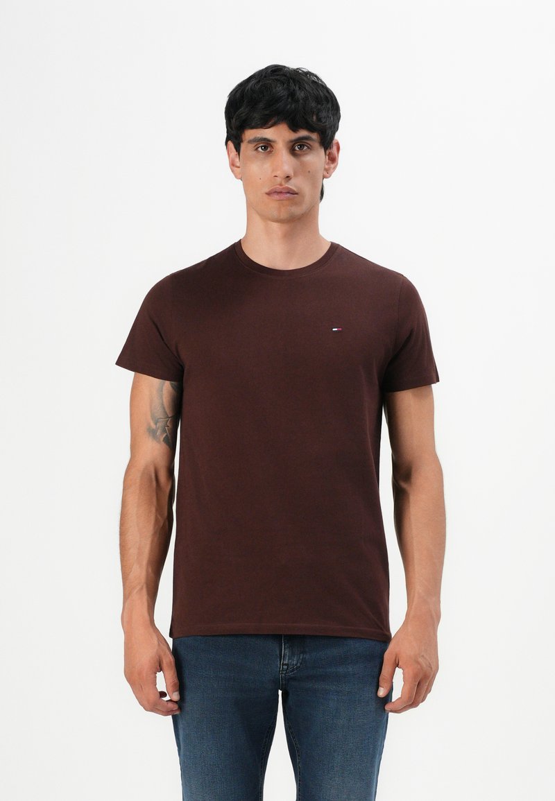 Ein Mann mit kurzen schwarzen Haaren trägt ein einfaches dunkelbraunes T-Shirt und blaue Jeans und steht vor einem weißen Hintergrund mit einem neutralen Ausdruck.