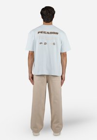 Light blue short-sleeve t-shirt featuring "PEGADOR" in bold brown letters on the back, with additional branding and graphic details below.