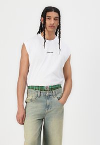 Hvitt ermeløst T-skjorte med svart logo, parret med lys blå denimjeans med grønn rutete midje. Uformell passform og avslappet stil.