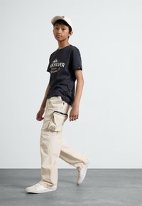 Svart t-skjorte med kort erme og logo, beige cargo-bukser med glidelåslomme, og beige sneakers, båret med en lysfarget caps.