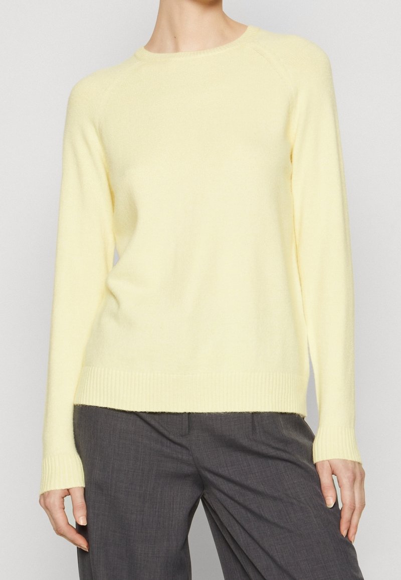 Personne portant un pull en maille jaune pâle à manches longues associé à un pantalon ajusté gris foncé, debout contre un fond clair uni.