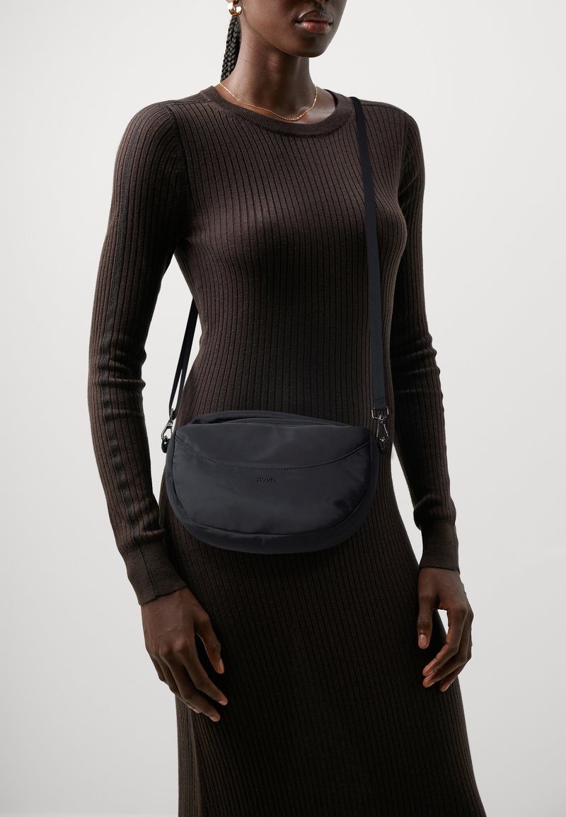 Sac banane en nylon noir avec une forme courbée et une fermeture éclair. Doté d'une bandoulière réglable et d'un logo en relief. Porté avec une robe marron côtelée.