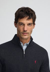 Dunkelgrauer Reißverschluss-Pullover mit geripptem Kragen und kleinem rotem Logo. Darunter ist ein hellblaues Hemd mit Kragen sichtbar.