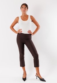 Canotta bianca aderente abbinata a pantaloni cropped marrone scuro e décolleté nere a punta. La modella posa con le mani sui fianchi contro uno sfondo bianco.