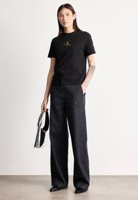 Jeune femme avec un bras tatoué portant un t-shirt noir CK et un pantalon large foncé, tenant un sac à main argenté, debout devant un fond uni.